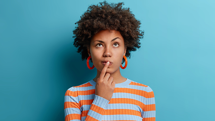 Vertical Shot Of Thoughtful Dark Skinned Woman Has Idea In Mind, Keeps Finger On Lips, Looks Pensively Above, Tries To Decide How To Act, Poses Against Blue Background, Blank Copy Space Above