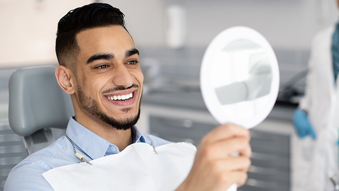 Happy Handsome Arab Man Looking To Mirror After Teeth Treatment In Clinic
